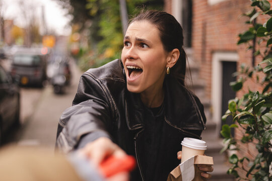 Scared young brunette woman in black leather jacket shouting in terror as thief snatches red smartphone from her hand on city street, frightened female victim holding coffee and paper food bag