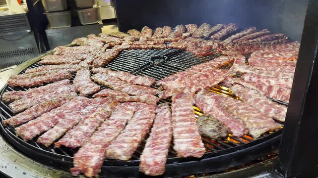 Pork ribs rotate on a metal grill slowly roasting at restaurant kitchen. The meat sizzles over open heat developing rich golden crust. The focus on cooking process, texture and appetizing appearance