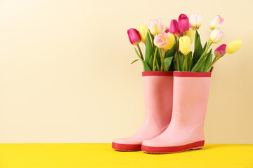 Fototapeta premium Beautiful tulip flowers in pink rubber boots on yellow wooden table against beige background. Space for text