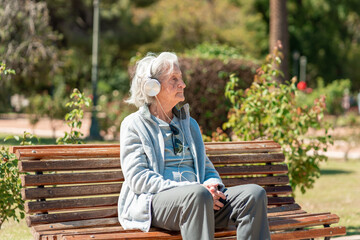 Fototapeta premium An 85 year old grandmother enjoys music with headphones while sitting on a park bench