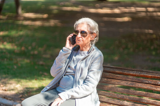 An 85 year old grandmother talks on her phone while sitting on a park bench with her daughter