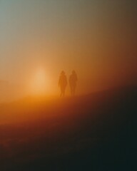 Fototapeta premium Two silhouetted figures walking through a foggy landscape at sunset, with warm orange and blue hues creating a cinematic film still aesthetic