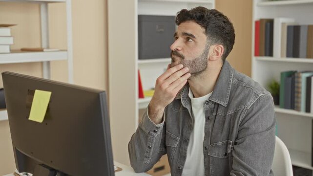 Man touches chin and types at desktop computer monitor in building office with a sticky note on the screen, gentle smile; focused productivity.