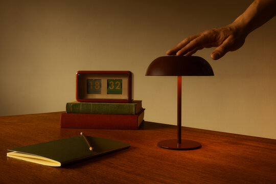 A hand reaching for a retro mushroom lamp on a wooden desk with a vintage flip clock, old books, and a notebook. Warm, moody lighting creates a cozy, nostalgic mid-century modern aesthetic workspace.