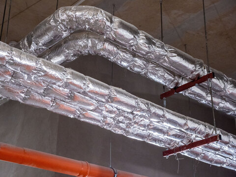 Industrial air ducts with silver thermal insulation, orange pipe on ceiling. Large air ventilation ducts are wrapped in shiny reflective silver thermal insulation foil, suspended from concrete ceiling