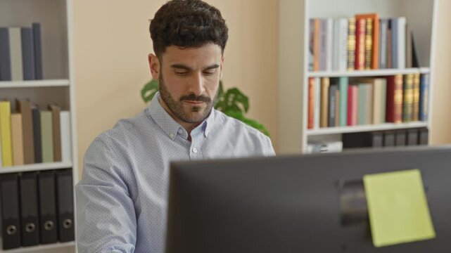 Man rubbing neck and wincing while seated at a computer desk with bookshelves and binders visible in an office building; work fatigue.