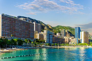 Fototapeta premium scenic cityscape of embarkment of Monte Carlo city in Monaco with blue sea water bay and amazing tall buildungs and houses along the coast. Urban mideterranean landscape with mountains and cloudy sky.