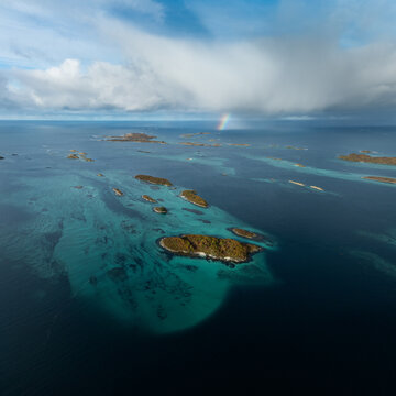 Aerial view of small rocky islands scattered across turquoise and deep blue waters under a cloudy sky with a faint rainbow near Hamn i Senja in Skaland, Troms, Norway.