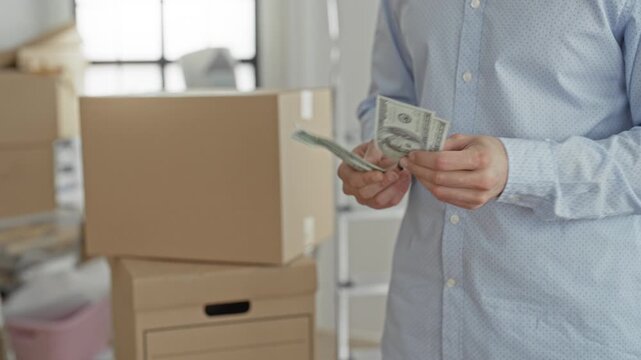 Man counting dollar bills with hands beside stacked cardboard boxes and moving supplies in a building during a move; budget anxiety.