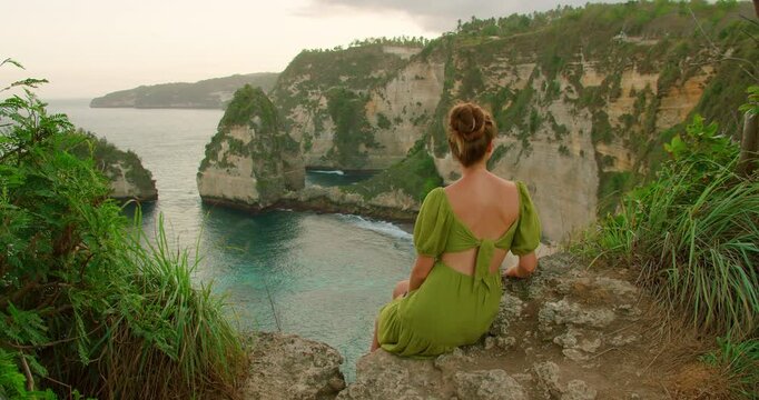 Back view of woman sitting on cliff overlooking blue ocean and Dimond beach in Nusa Penida Island Bali