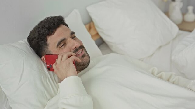 Man lying in bed holding smartphone to ear, smiling and gesturing with hand in a bedroom; relaxed conversation.
