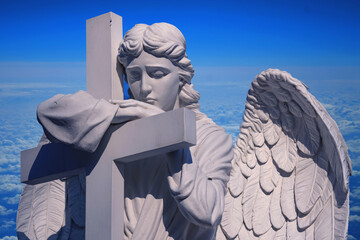 Fototapeta premium Angel statue leaning on cross above clouds, symbol of faith, hope and eternal life.