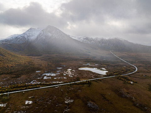 Aerial view of Ballesvika valley featuring a winding road, snow-capped mountain peaks, and autumn tundra under a dramatic cloudy sky in Ballesvika, Troms, Norway.