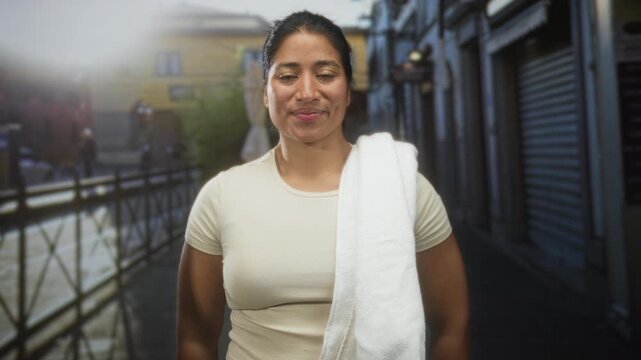 Young sporty woman with towel over shoulder smiles with eyes closed and waves hand on an urban street with shuttered shops; post workout confidence.