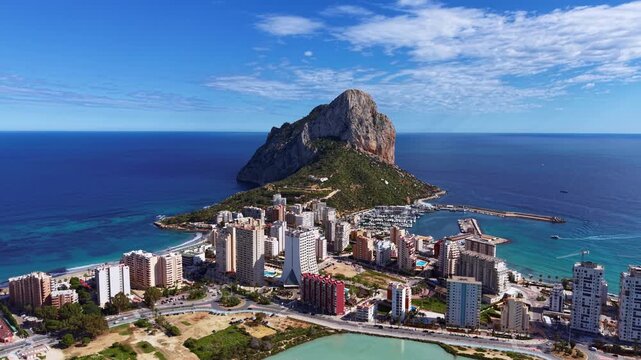 Aerial panoramic view of Calpe, Spain, featuring the iconic Penyal de Ifac rock formation rising above the coastal city, turquoise Mediterranean waters, marina and salt lagoon
