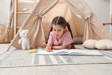 Little girl drawing in sketchbook near toy wigwam at home © New Africa
