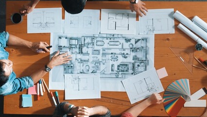 Top down aerial view of professional project manager planing house design by using plan. Aerial view of smart team working together discussing while pointing at blueprint at meeting table. Symposium. © InfiniteFlow
