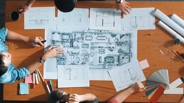 Top down aerial view of professional project manager planing house design by using plan. Aerial view of smart team working together discussing while pointing at blueprint at meeting table. Symposium.