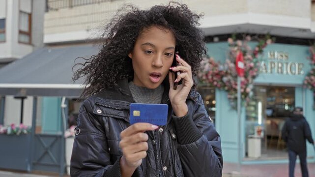 Woman holding a blue creditcard in her hand while talking on a smartphone outside a storefront on a busy street, looking at the card and listening intently; concern payment problem.