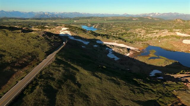 Aerial footage captures Beartooth Highway in Montana winding through alpine terrain with alpine lakes and melting snow patches. Distant snow-capped peaks frame the vast wilderness landscape.