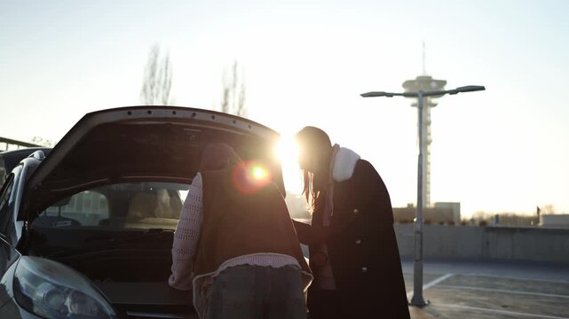 Sunlit scene two women stand by open car hood experimenting with tools and intuition trying to revive silent engine turning breakdown into unexpected learning moment in parking lot