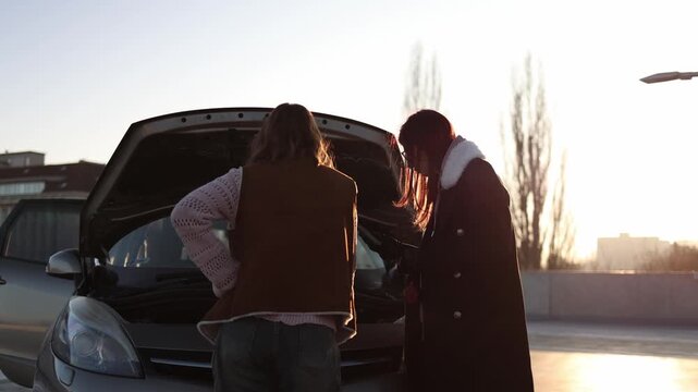 Two women stand by open car hood in sunlight checking engine together trying to fix breakdown following online advice learning repair for first time in parking lot