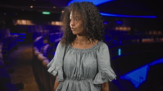 Teenage woman with curly hair tilting head and looking down in a cinema, wearing a ruffle dress; contemplation.