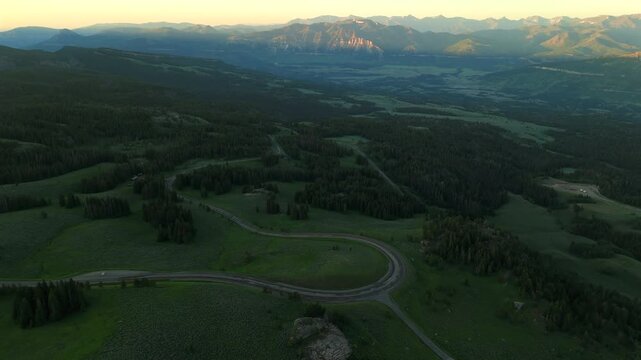 Aerial footage of Beartooth Highway in Montana showcases winding roads through lush green valleys with dense pine forests and distant mountain ranges bathed in golden sunset light.