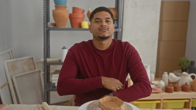 Man with clay on a pottery wheel and arms crossed in studio, beard visible, seated at pottery table; confidence craft focus.