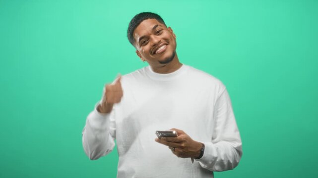 Man holding smartphone with thumb up while smiling and browsing on device against green studio backdrop wearing white long sleeve top; playful joy.