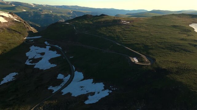 Beartooth Highway in Montana curves dramatically through high alpine terrain with patches of melting snow dotting green meadows. Golden hour light illuminates the scenic mountain pass landscape.