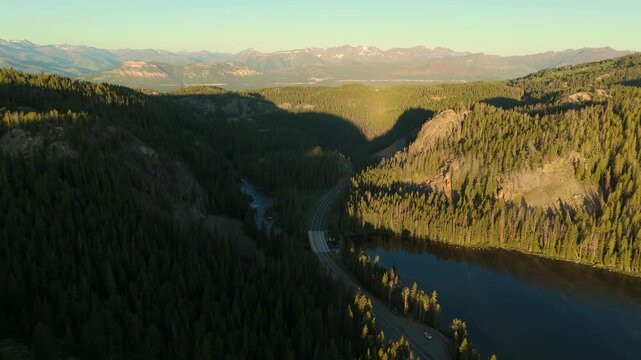 Beartooth Highway in Montana curves through a scenic mountain valley with dense evergreen forests and a tranquil lake. Snow-capped peaks rise in the distance under golden hour light.