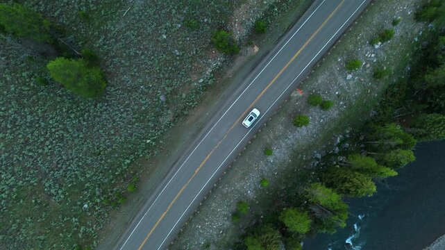Drone footage captures a white car traveling along the scenic Beartooth Highway in Montana. The winding mountain road cuts through rugged rocky terrain and lush green forest near a river.