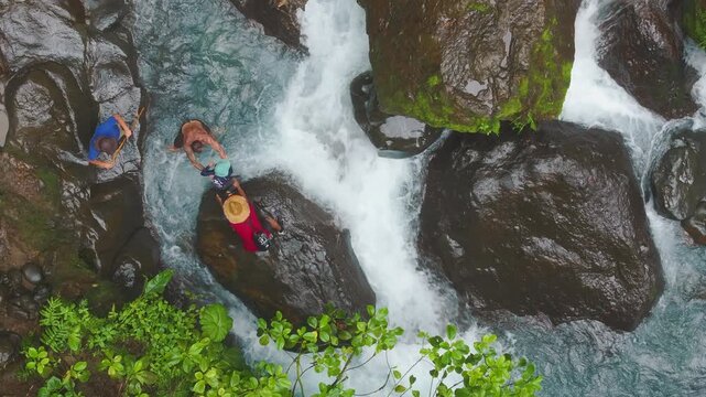 Top down aerial view of three men passing a young boy hand to hand while crossing a rocky river in tropical forest, teamwork and support concept.