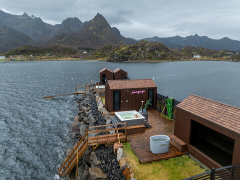 Aerial view of Skarungen spa featuring wooden cabins, a hot tub, and a cold plunge pool on a deck by the water with rugged mountains in Kabelvag, Nordland, Norway.