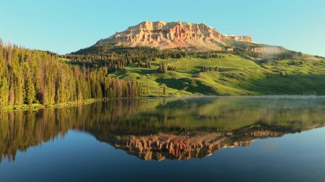 Stunning mountain peak with red rock cliffs reflects perfectly in glass-like alpine lake water. Morning mist rises over evergreen forest as golden sunrise light illuminates the rugged landscape.