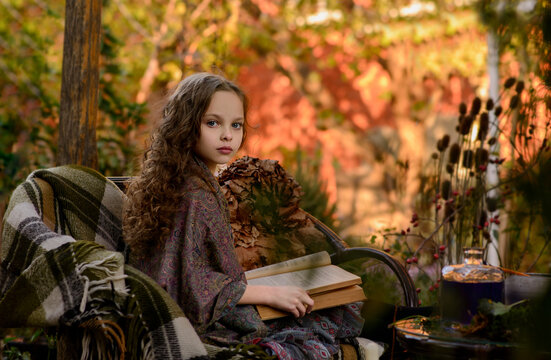 Cute little girl with long curly hair reads big old book sitting on the bench outdoor in the autumn garden. Beautiful child girl is reading retro book. Halloween and autumn season.Selective focus
