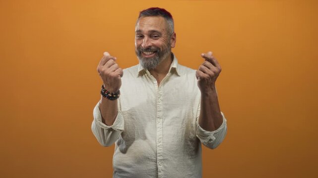 Man middle aged with grey hair and beard smiling, hands pinching fingers wearing beaded bracelets and linen shirt in orange studio; playful charm confidence.