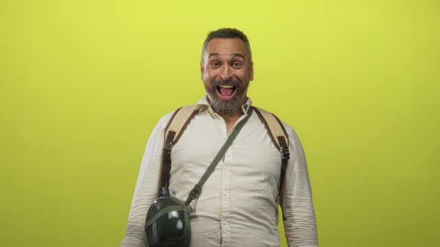 Man middle aged with grey hair and beard with backpack and canteen smiling and laughing in lime green studio; joyful adventure travel.