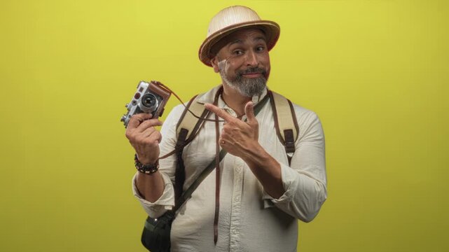 Man holding a vintage camera and pointing to it while wearing backpack and pith helmet in studio with bright yellow backdrop; adventurous traveler.