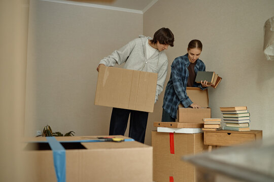 Young adult couple unpacking moving boxes and sorting books in new home, preparing relocation and fresh start. Useful for real estate, rental services, mortgage, moving company marketing