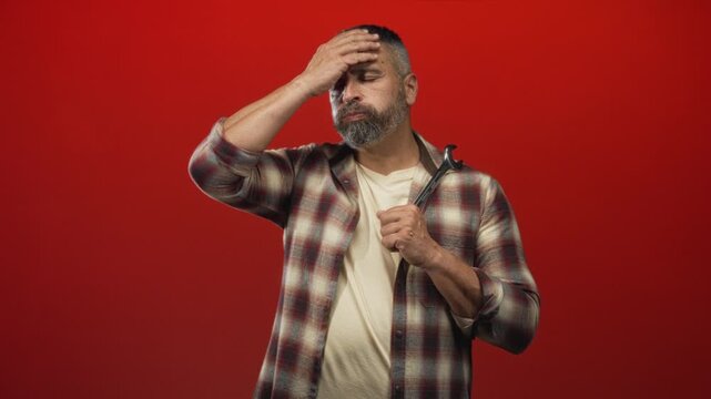 Man mechanic holding wrench and gripping shirt while pressing forehead with hand in red studio; fatigue resilience.
