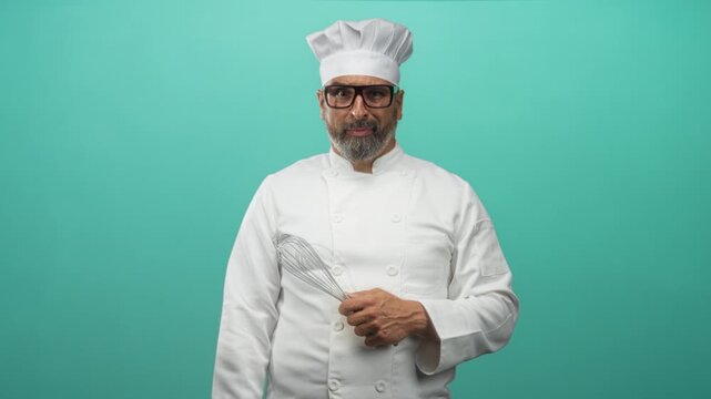 Man chef holding whisk and wearing chef uniform and hat in turquoise studio; confidence skill expertise.
