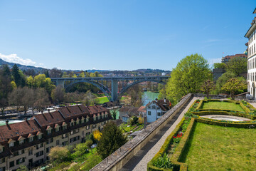 Obraz premium Panoramic spring cityscape of Bern with the Aare River, Kirchenfeld Bridge, old town rooftops and terraced gardens under a clear blue sky.