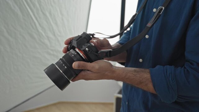 Man photographer holding camera and adjusting zoom lens with bare hands, strap visible, rolled sleeves and exposed forearms in studio softbox setup; concentration.