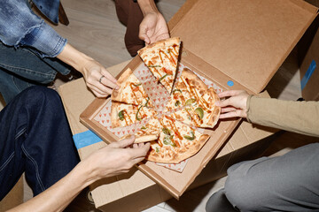Overhead shot of young adults sharing pizza from delivery box, grabbing slices during casual group...