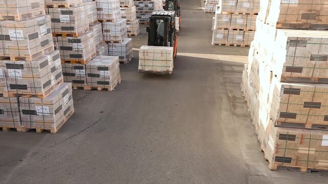 A forklift carries boxes through an outdoors warehouse. A forklift transports goods through an outdoors warehouse. Storage yard, warehouse yard. Finished factory products in boxes on pallets