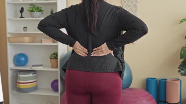 Woman with hands on lower back near exercise ball in studio wearing black top and burgundy leggings; backache discomfort.