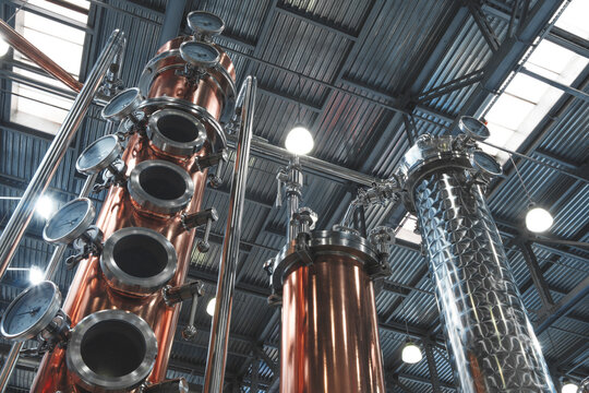 interior of an industrial distillery features tall copper and stainless steel columns. Pressure gauges and pipes indicate the ongoing distillation process. The high ceiling features exposed beams.