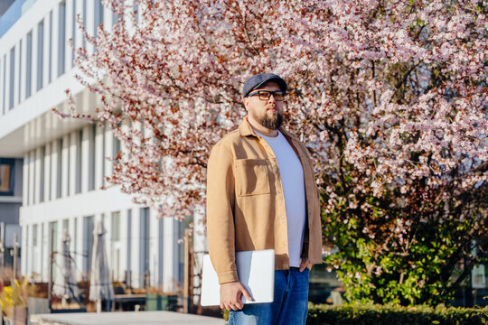 freelancer man holding laptop and standing in city near blooming cherry tree